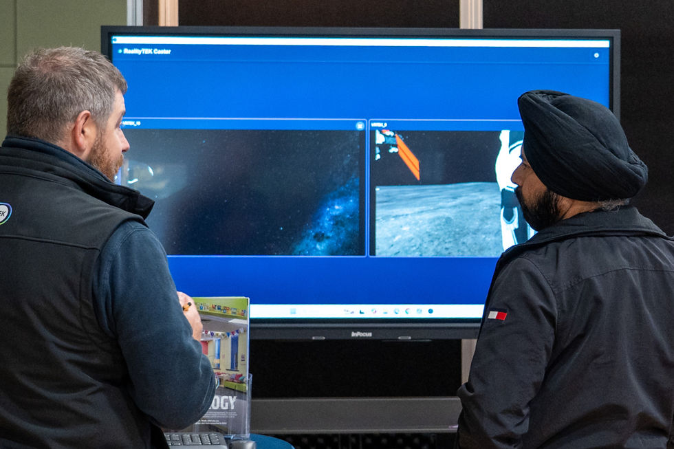 Two men discuss images on a large screen displaying space scenes. One holds a brochure. The mood is focused.
