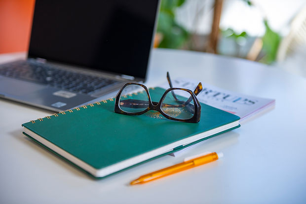 An open laptop, a closed notebook and a pair of glasses sit on a desk.