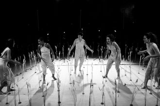 Seis atrizes em cena, em um cenário que remete a um campo de trigo. Elas se olham e cantam.