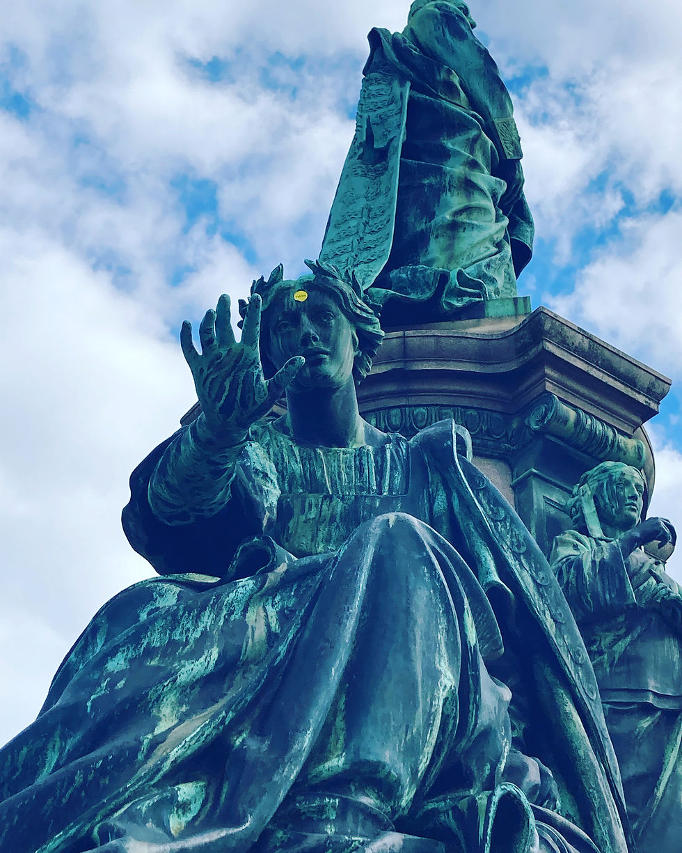 Statue with hand towards the camera has a bright yellow sticker against the green copper on her forehead. In front of a blue and clouds sky. 