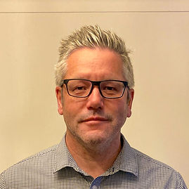 Image of Jonathan McCrea, a white man standing centre frame, he is wearing black glasses, and a checkered shirt, he has short gret hair