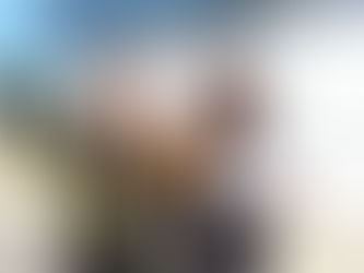 Two people smile outdoors; one holds orange roses and a certificate. Sunlit building in the background. Celebratory mood.