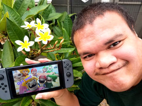A person smiles holding a gaming console displaying cartoon characters and "Get well Number 7!". White flowers and green leaves in the background.