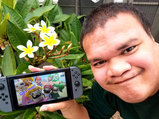 A person smiles holding a gaming console displaying cartoon characters and "Get well Number 7!". White flowers and green leaves in the background.