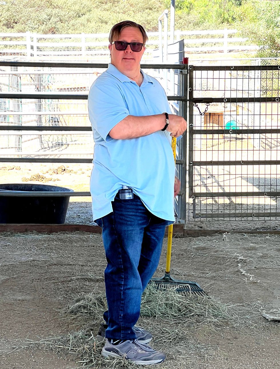 A person with Down syndrome in sunglasses, light blue shirt, and jeans holds a rake in a sunny, fenced outdoor area with hay and trees in the background.
