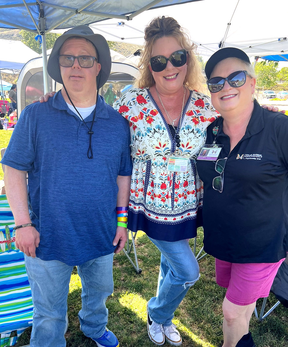 Three people smiling under tents at an outdoor event. One wears a hat and blue shirt; others in sunglasses and colorful outfits. Sunny day.