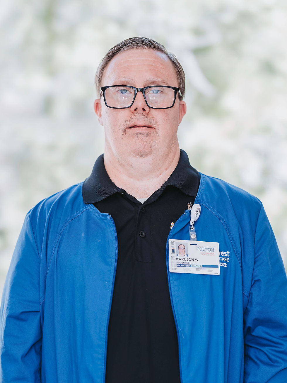 A person with Down syndrome in a blue jacket with a volunteer badge stands against a blurred background. They look directly at the camera with a neutral expression.