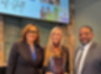 Three people smiling indoors. Woman in center holds an "Award of Appreciation" plaque. Background shows a wall with photos and text.