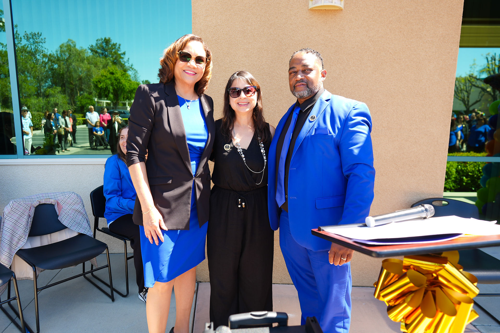 Three people standing together smiling in front of a building. One wears a blue dress and black blazer, another wears a black outfit, and the third wears a blue suit.