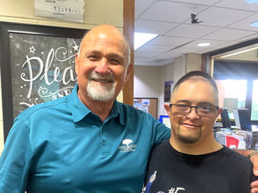 Two smiling men, one with Down syndrome, pose together in an office. One wears a blue shirt, the other a Dodgers MVP T-shirt. A sign reads "Please" in the background.