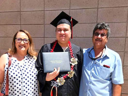Graduate with autism in cap and gown holding a diploma, flanked by smiling woman in polka dot shirt and man in blue shirt, against brick wall.