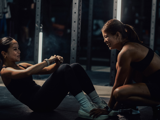 Two women in a gym, one doing sit-ups and the other supporting her feet, both smiling. Dark setting, bright lights in the background.