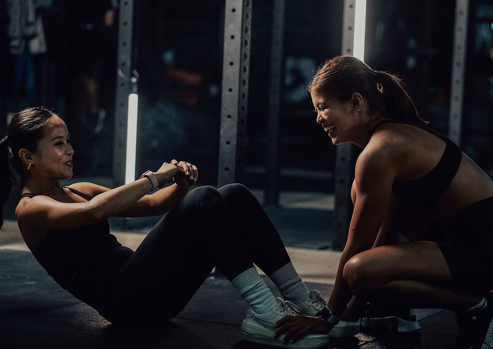 Two women in a gym, one doing sit-ups and the other supporting her feet, both smiling. Dark setting, bright lights in the background.