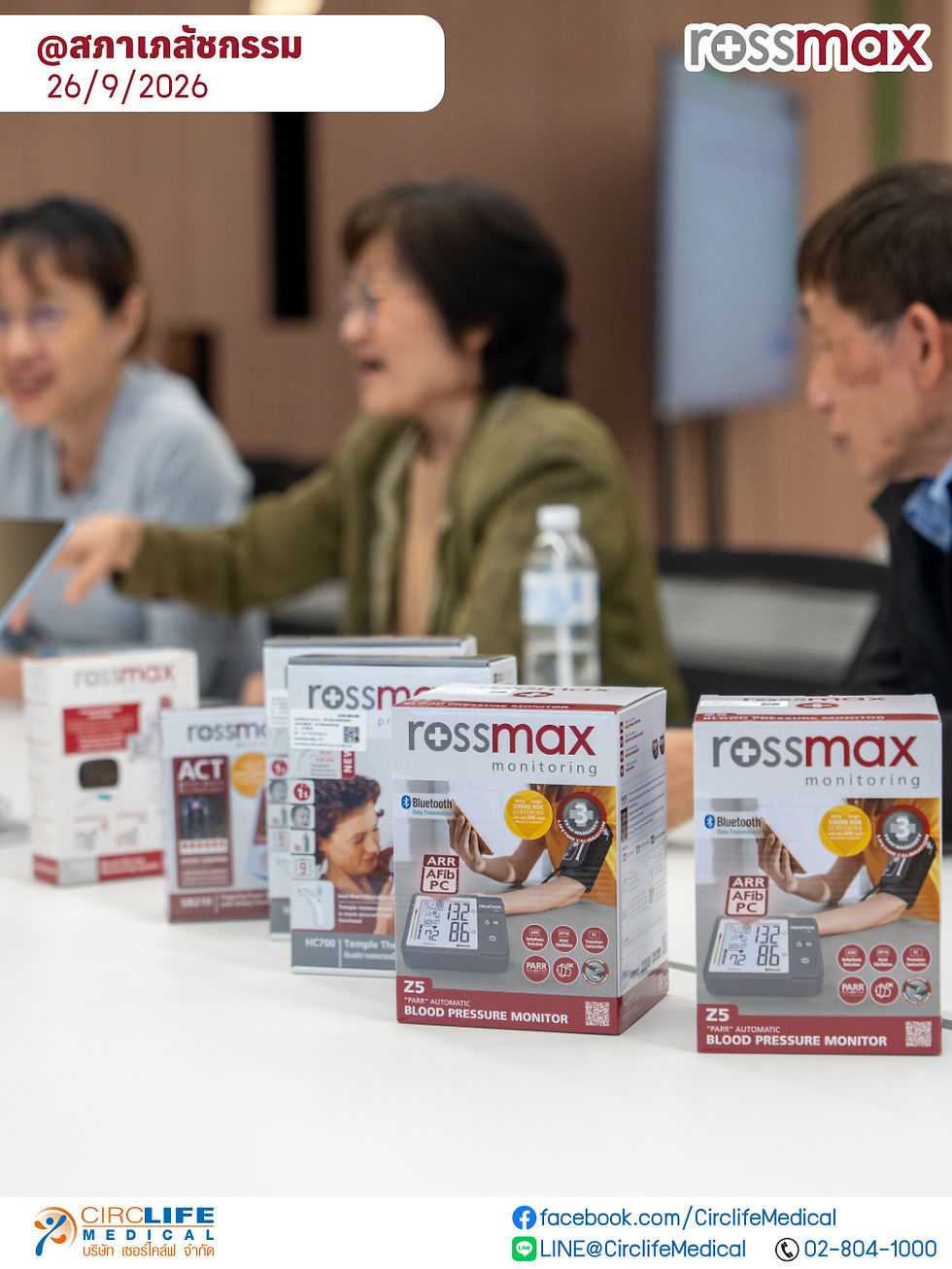 Blurred meeting with three people behind rows of Rossmax blood pressure monitor boxes; Thai date @Pharmacy Council 26/9/2026.