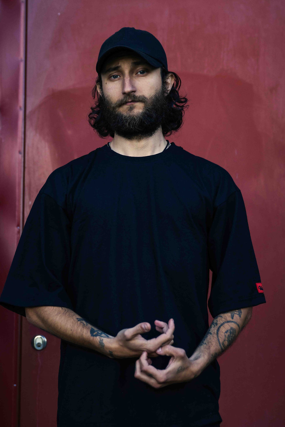 Thumbnail: Man in a black oversized t-shirt with a minimalist look, standing against a red door. He has long hair, a beard, and tattoos