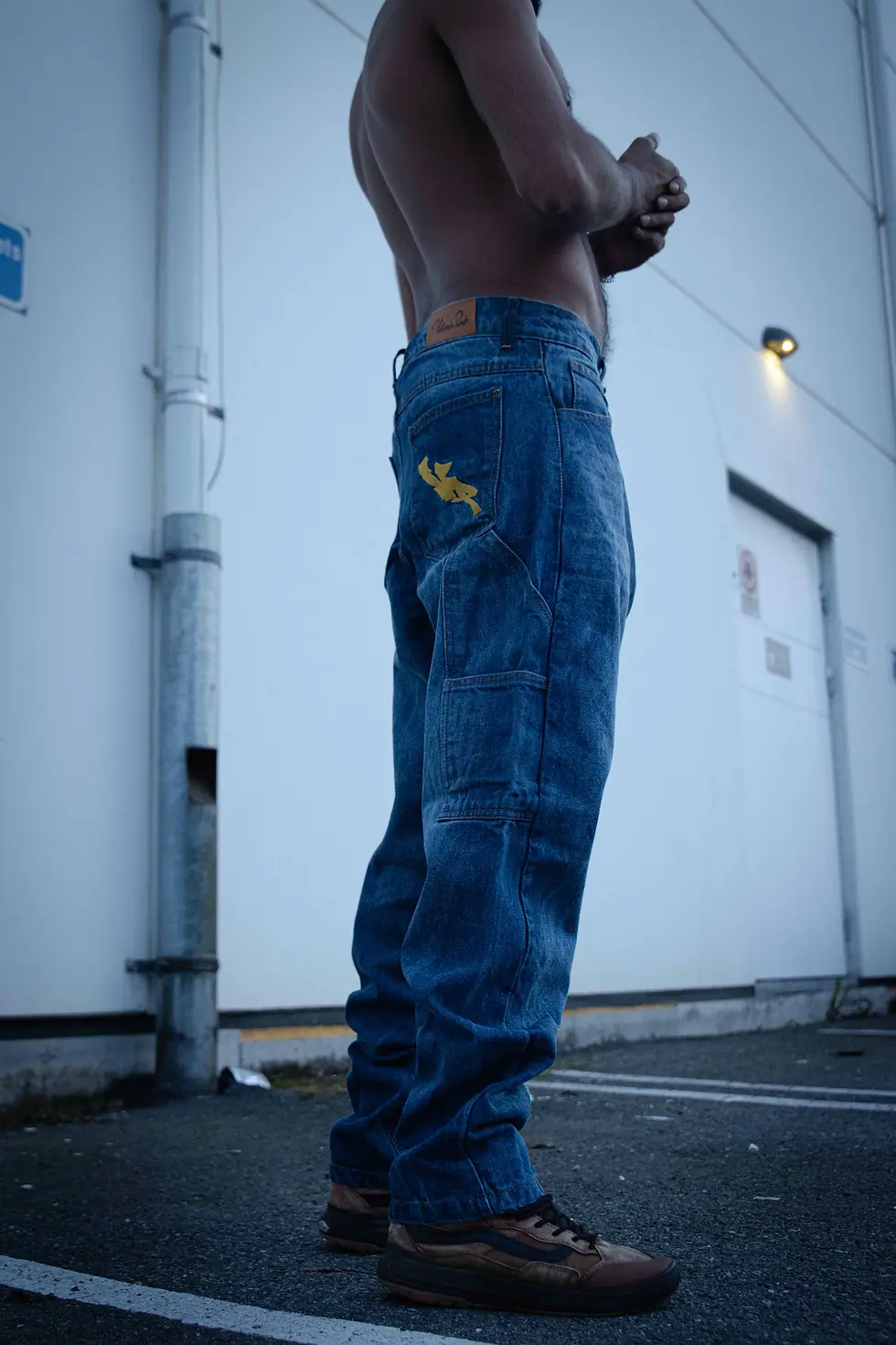 Thumbnail: Side view of a person in baggy blue jeans, with arms crossed, standing near a building.