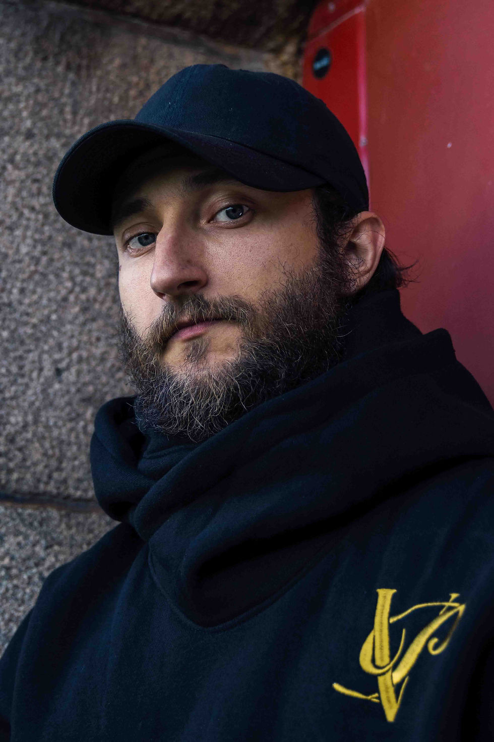 Thumbnail: Portrait of a person wearing a black hoodie with a yellow logo, standing against a stone wall.