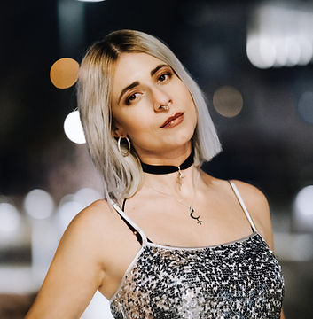 Junge Frau mit kurzen blonden Haaren und einem silbernen Glitzertop. Sie schaut in die Kamera, die Hand in die Hüfte gestemmt.