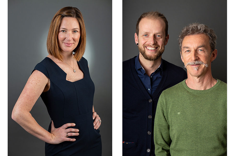 A woman in a dark navy dress smiles confidently, hands on hips. Two men in sweaters stand side by side, smiling against a grey background. Both photographed by Andrea Russell