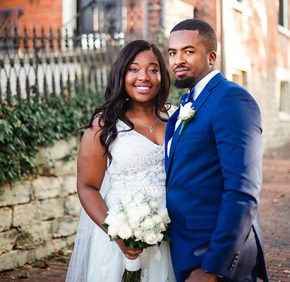 A woman in a wedding dress and holding a bouquet of white roses, and a man in a navy blue suit, looking to the camera