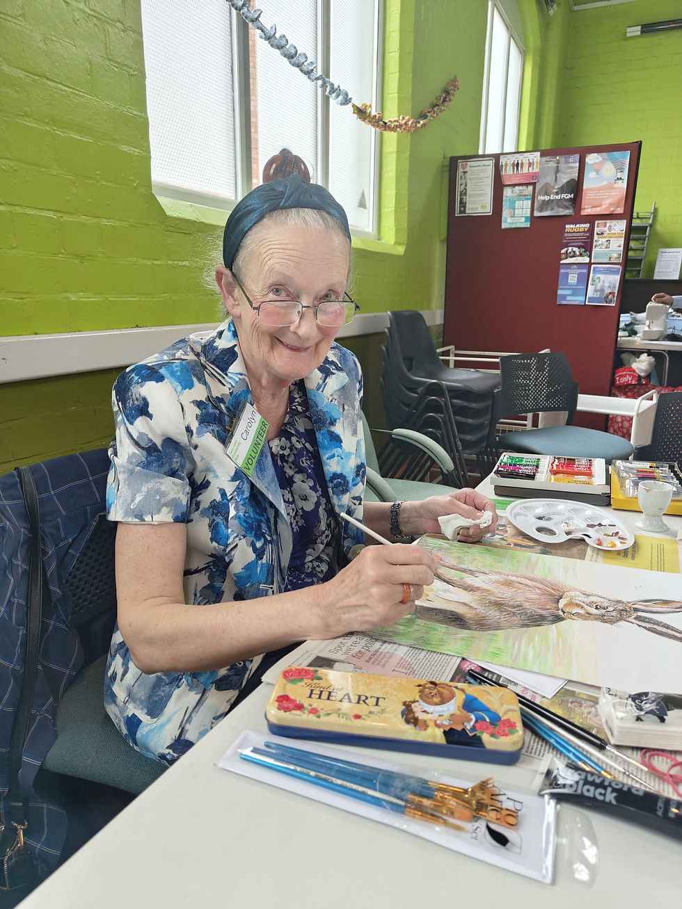 Photo of Carolyn, smiling with glasses and wearing a blue flowery shirt, painting a rabbit at Wednesday Warm Welcome
