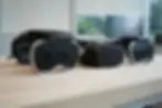 Meta VR headsets on a wooden table in an office setting. Background shows blurred screens and greenery outside a window. Black with white accents.