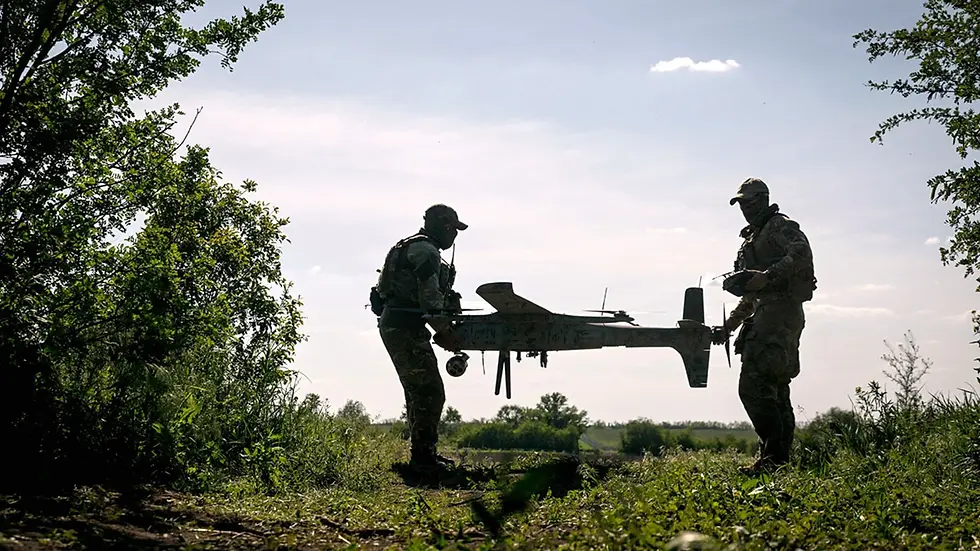 Two soldiers in silhouette handle a large drone in a grassy, wooded area. The sky is bright with scattered clouds, creating a tense mood.