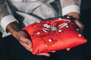 Hands hold a red embroidered ring pillow with wedding bands, suggesting a formal wedding moment.