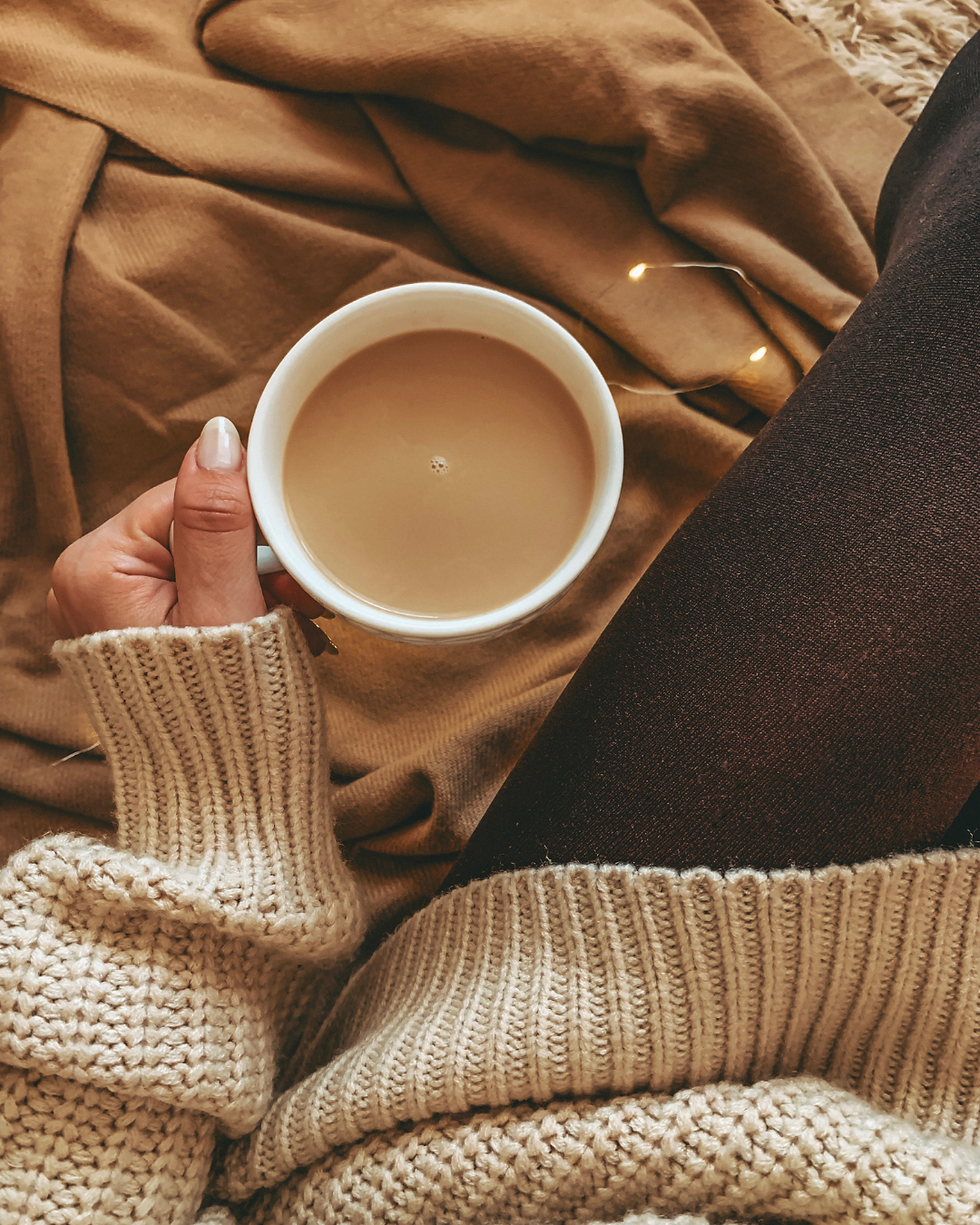 Hand holding a mug of coffee on a cozy tan blanket, with cream knit sweater and dark tights in warm light.