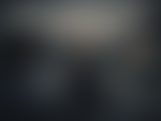 Two women speaking in a suburban driveway at dusk while a faint human figure stands near a house in the background, illustrating a subtle paranormal encounter.