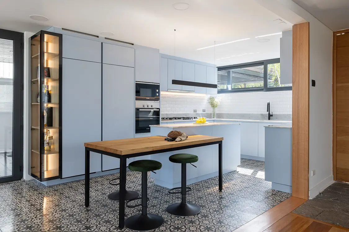 Cocina moderna minimalista en azul pastel con muebles sin tiradores, combinada con un original suelo hidráulico de patrón geométrico. El diseño incluye una barra de desayuno de madera natural y metal negro, iluminación LED integrada en vitrina y electrodomésticos panelados. Ideal para inspirar proyectos de diseño de interiores luminosos y funcionales.