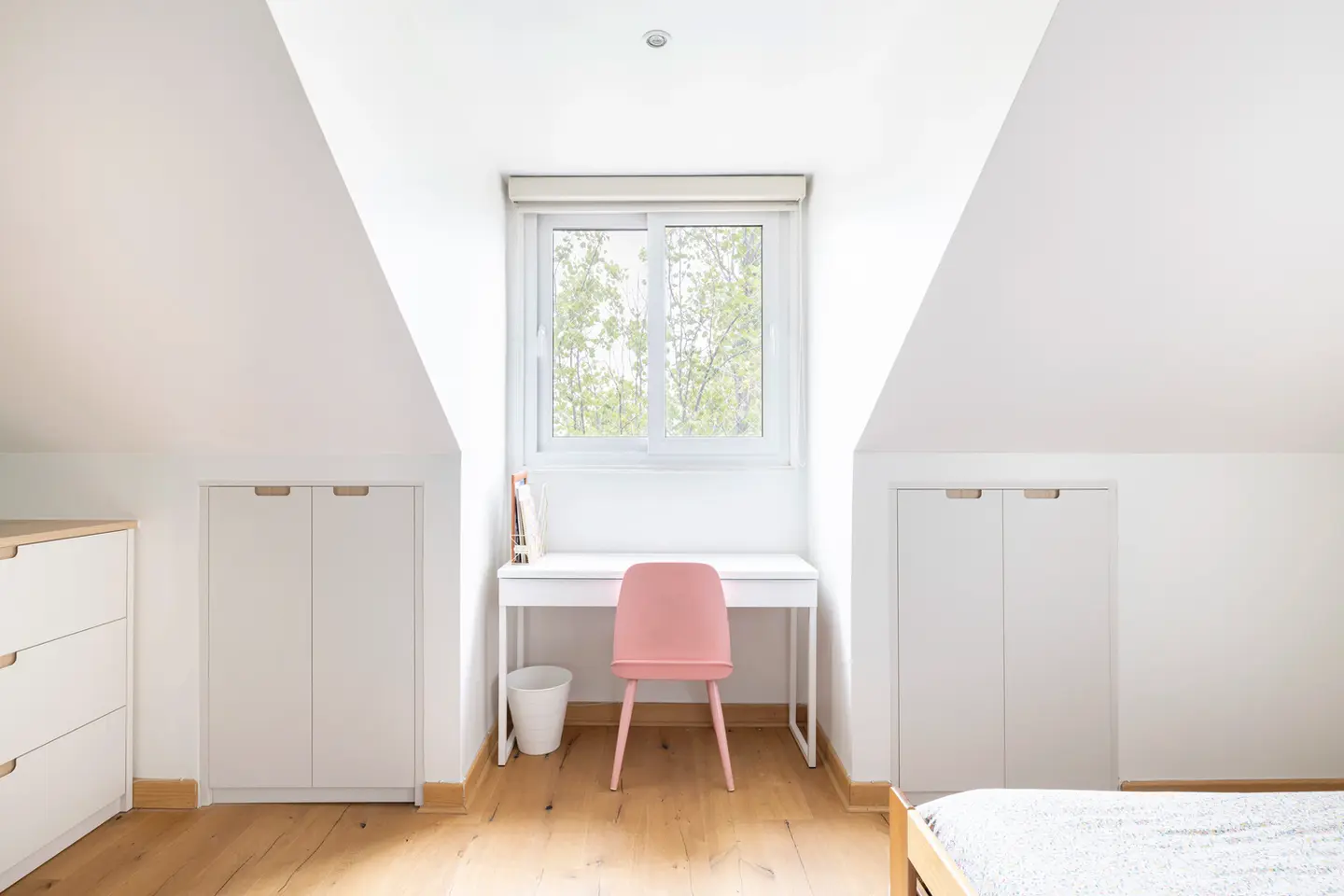 Escritorio blanco minimalista con silla rosada junto a ventana y armarios laterales en habitación abuhardillada