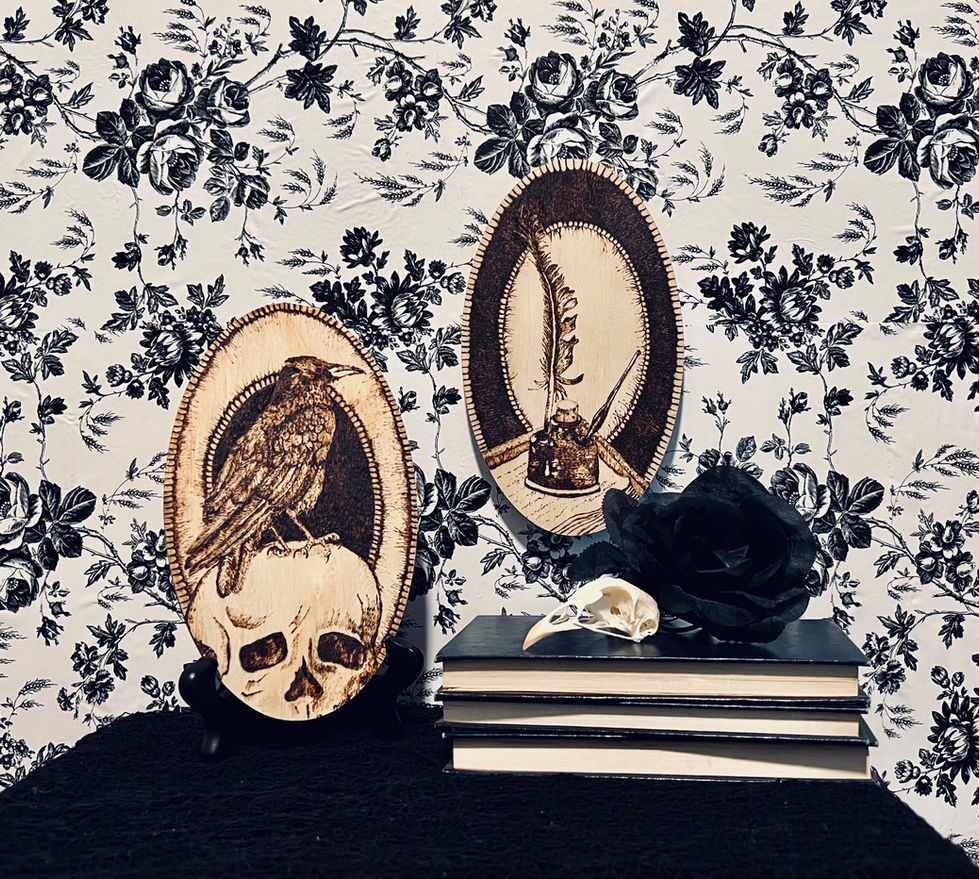 Artwork shown on a dark tabletop with black and white floral wallpaper behind. The piece with the skull and raven is propped against the wall, next to a stack of books. The inkwell piece hangs on the wall above the books.