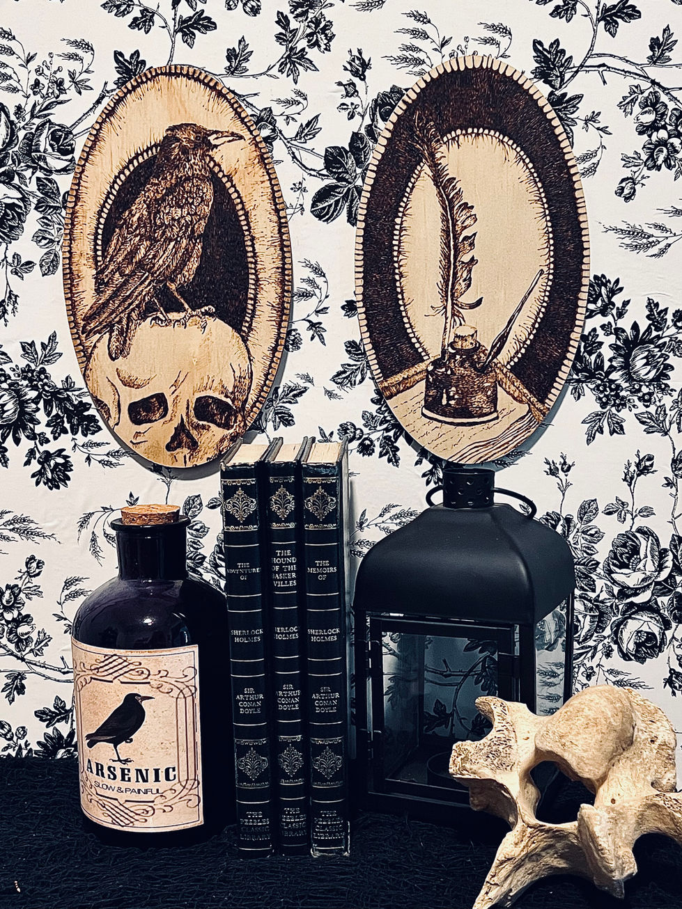 Art work hangs on a wall with black and white floral wallpaper. On the table below are several objects: a dark bottle labeled "arsenic," three black books with gold writing on the spines, a small black lantern, and a vertebrae bone from a cow.