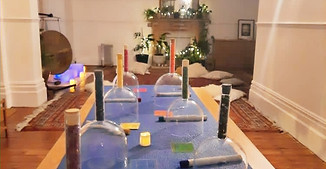 A set of 7 handheld crystal bowls, each handle filled with crystals the colour of a chakra. The set resting on a table in a studio ready for a sound bath. A gong and other white crystle bowls are in view.