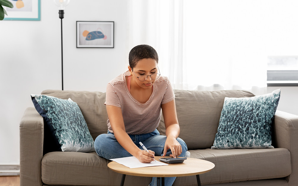 Female with short hair earing glasses sitting on sofa writing and using a calculator