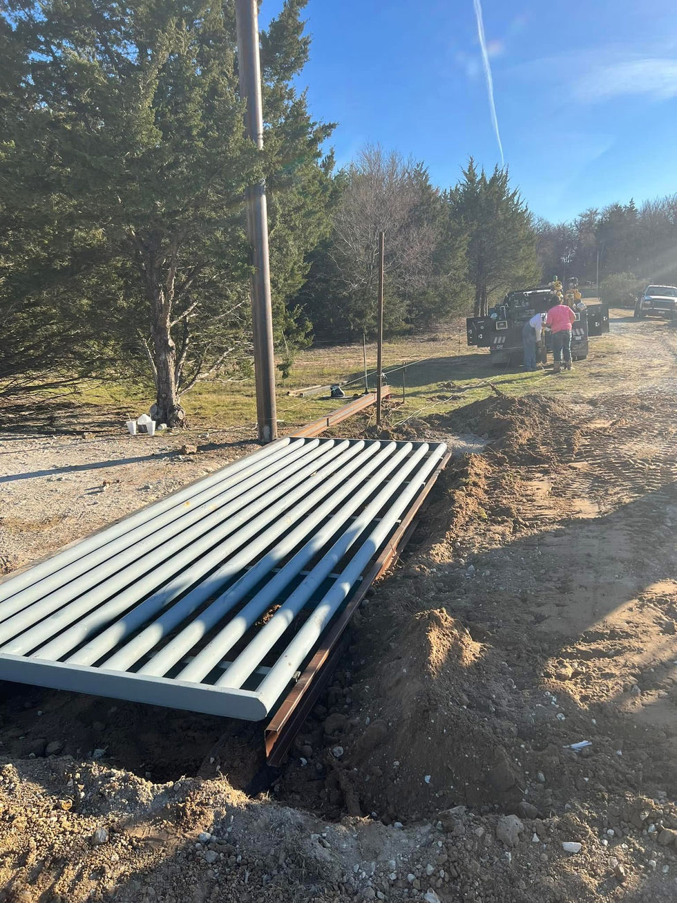 A metal cattle guard, crafted through custom fabrication, is being installed in a dirt road. Two people stand near a utility pole and a truck, with trees and grassy area in the background under a clear blue sky.