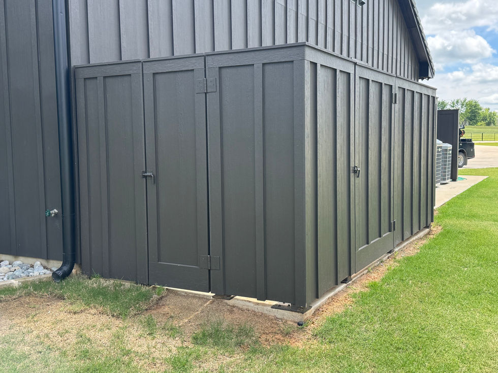 A dark gray wooden equipment enclosure with double doors is attached to the side of a building, set on a lawn with some bare patches near the base and a partly cloudy sky in the background.