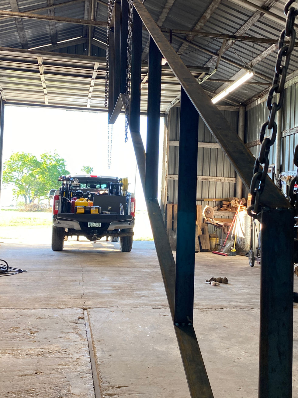 A metal structure and hanging chains inside a workshop hint at custom fabrication, leading toward an open garage door where a pickup truck loaded with tools is parked, with sunlight and greenery outside—ready for residential construction projects.