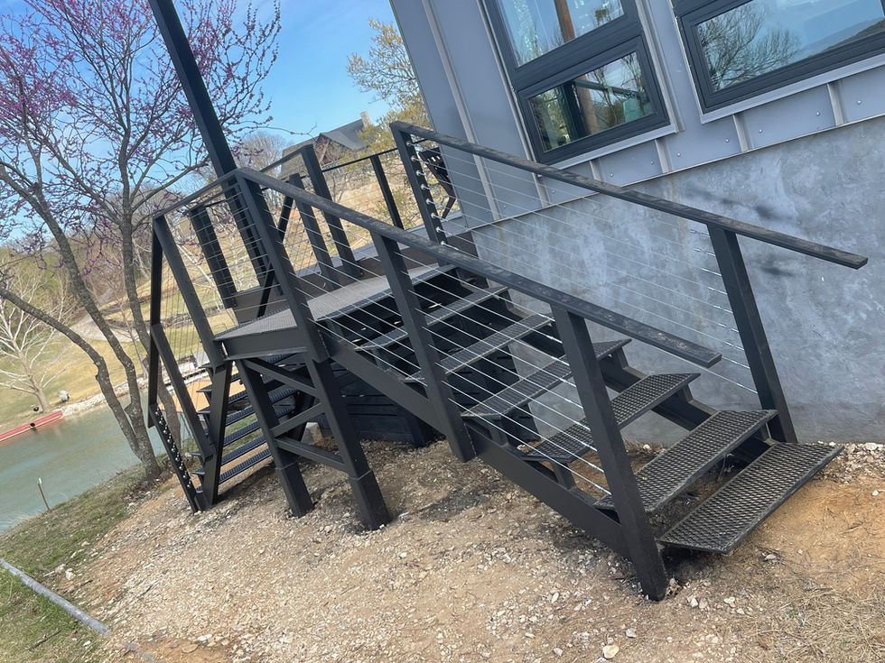 Black metal outdoor stairs with cable railing, custom fabricated for residential construction, lead up to a modern building with gray siding and large windows, set above rocky ground near a body of water and trees.