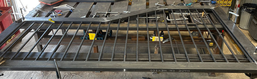 A large metal gate with vertical bars is being assembled on a workbench. Clamps, measuring tools, and other workshop equipment surround the custom gate, highlighting its mid-construction stage as part of a mobile welding project.