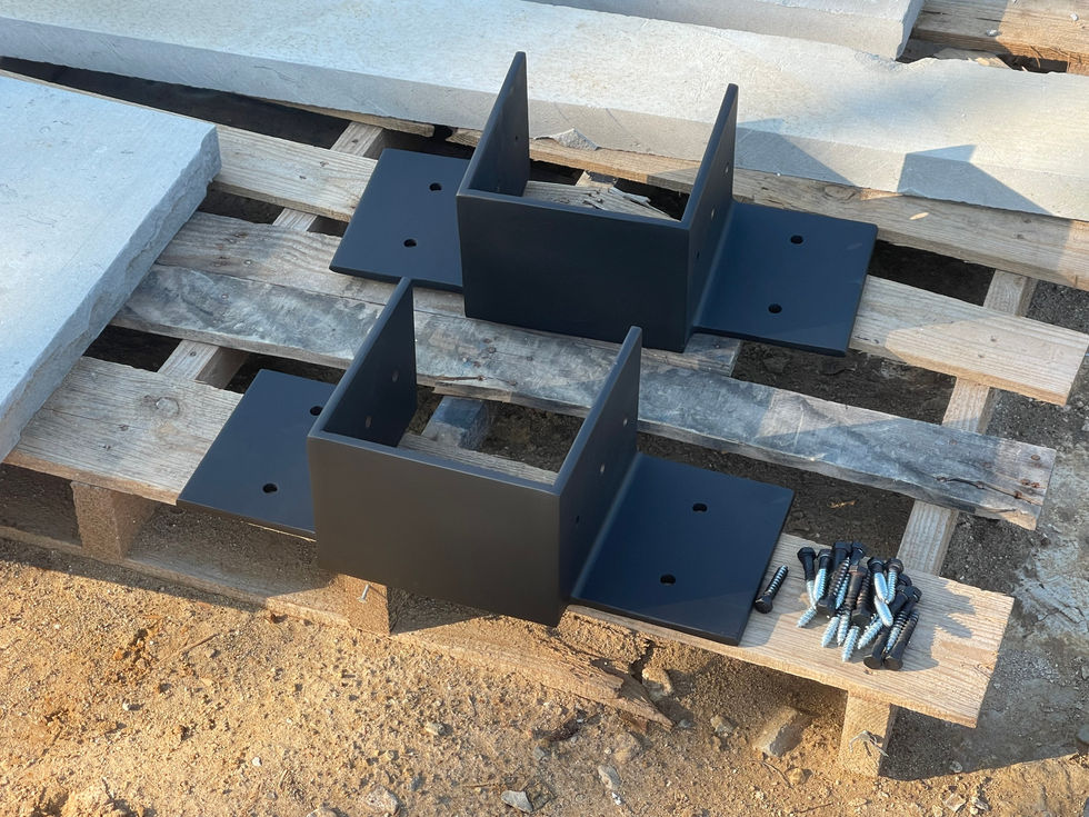 Two black industrial style brackets with mounting holes rest on a wooden pallet outdoors, screws nearby. Custom fabrication is evident, with concrete slabs stacked in the background.