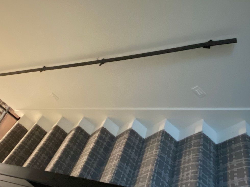 A black curtain rod, reminiscent of custom fabrication, is mounted high on a white wall above patterned curtains with a geometric design in grey and white. The photo is taken from a low angle.