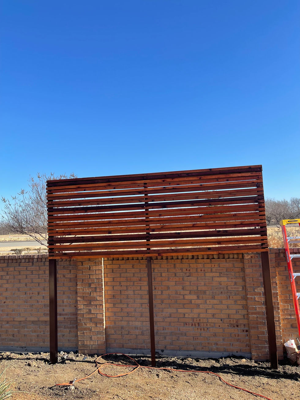 A wooden slat privacy wall is being built in front of a brick wall outdoors under a clear blue sky. A red ladder stands on the right, with construction tools on the ground, showing the first steps before mobile welding begins.