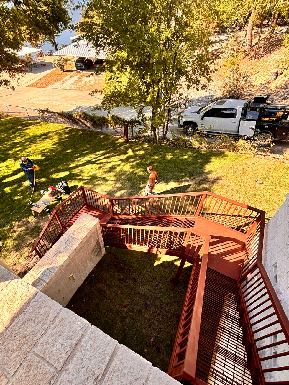 View from above of two people working in a grassy yard near a custom staircase attached to a white brick building, with tools and equipment scattered around and a white utility truck—ideal for mobile welding—parked nearby under trees.