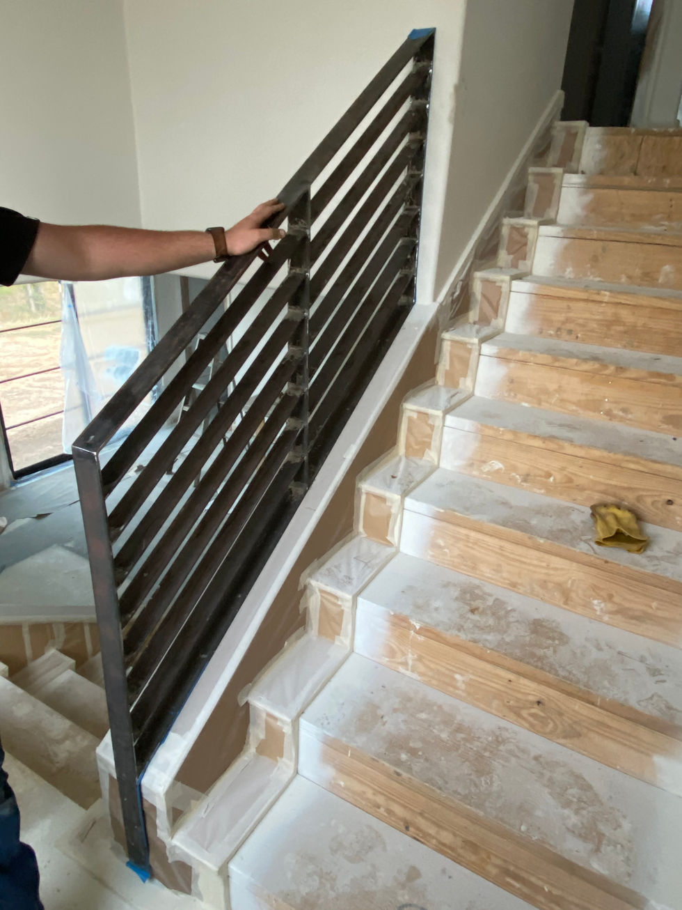 A person holds a custom railing made of black metal, installed on a staircase with unfinished wooden steps and white risers in a residential construction or renovation project.