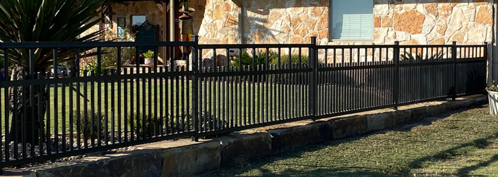 A single-story house with stone exterior walls, a brown shingled roof, two stone chimneys, a repiared metal fence perfect for custom fabrication or mobile welding, landscaped yard, palm tree, and blue sky in the background.