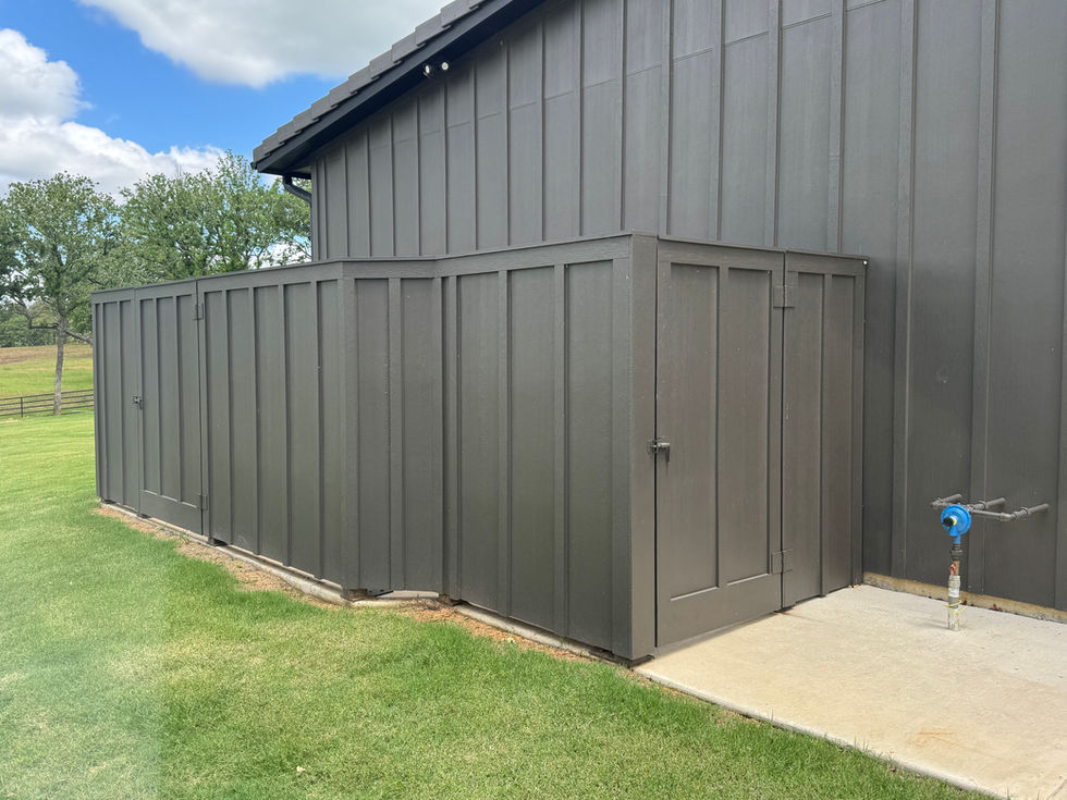 A dark gray wooden equipment enclosure is attached to the side of a building, featuring two doors and surrounded by green grass. Nearby, a water spigot and concrete pad offer space for mobile welding under a partly cloudy sky.