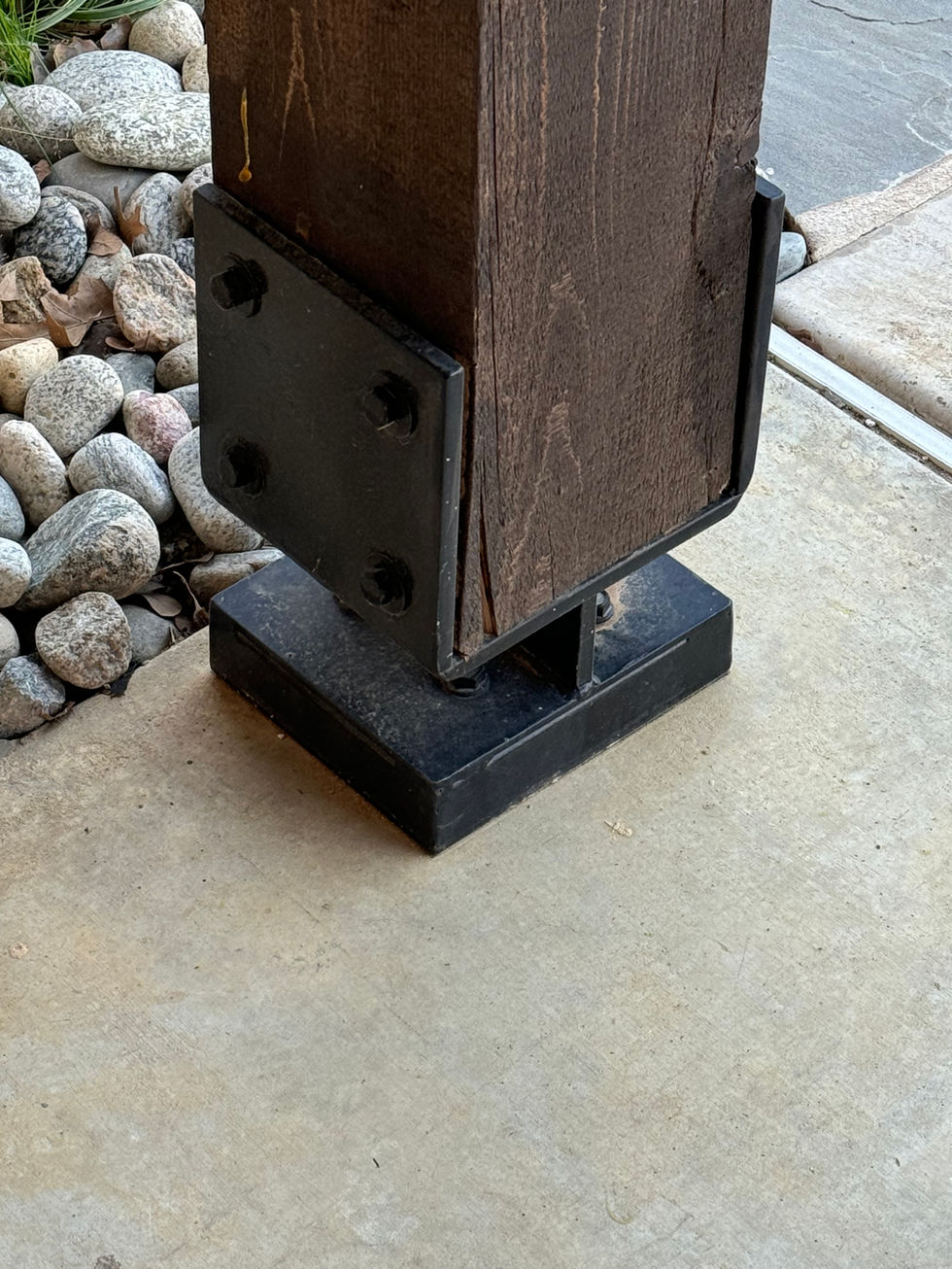 A wooden post secured to a concrete surface with custom fabrication industrial style brackets and bolts; decorative rocks are visible in the background.