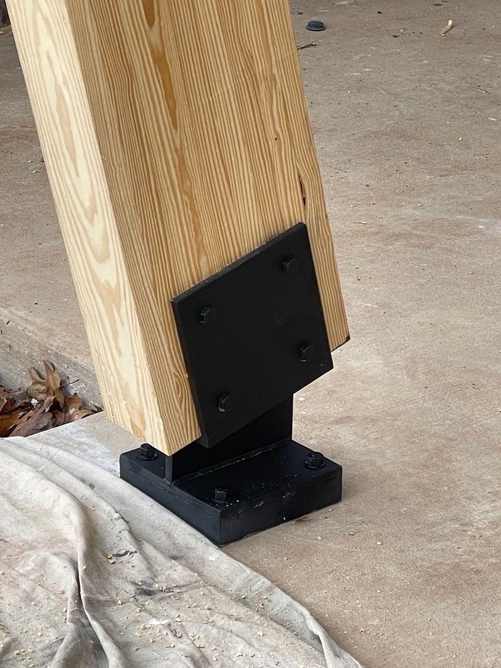 A wooden post is anchored to a concrete floor with a black metal base plate and bolts, showcasing custom fabrication. Nearby, a cloth or tarp is partially visible on the ground, surrounded by some sawdust and leaves.
